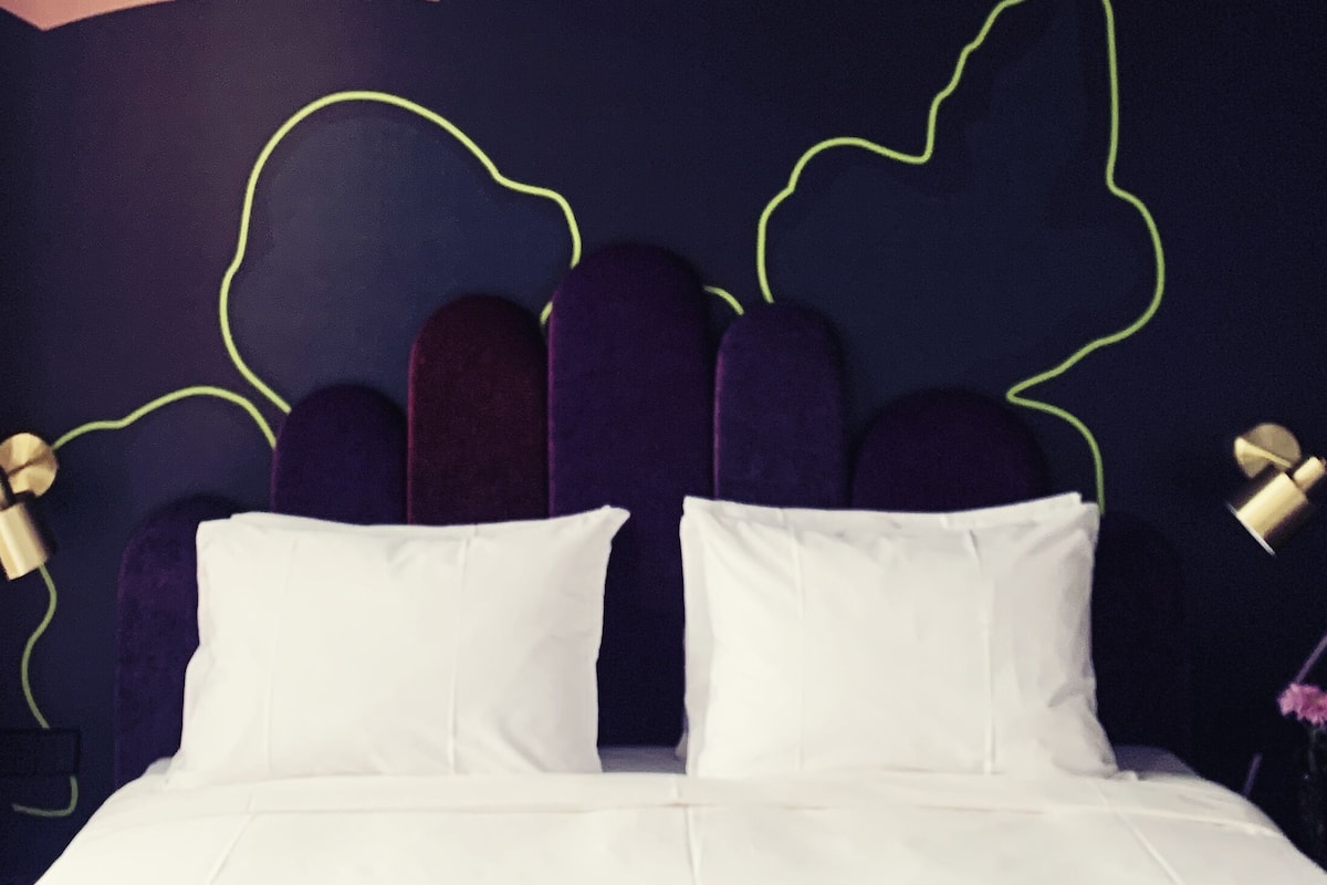 A bed with a white bedspread and two white pillows, a dark wall with neon green leaf outlines, and a partial view of a ceiling with a light fixture.