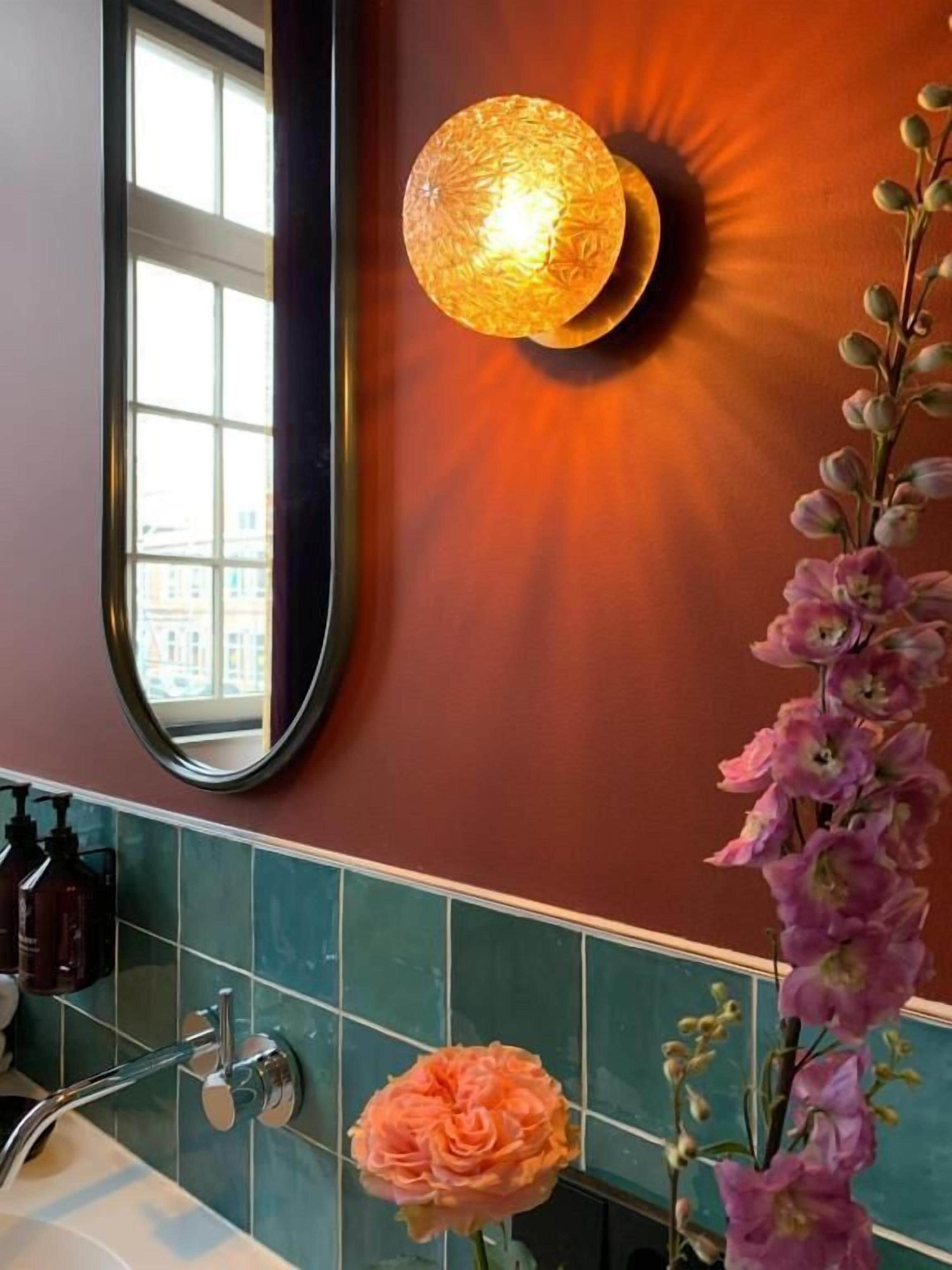 Un baño con espejo, una lámpara en la pared, un arreglo floral y una zona de ducha.