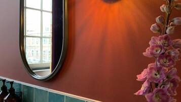 Un baño con espejo, una lámpara en la pared, un arreglo floral y una zona de ducha.
