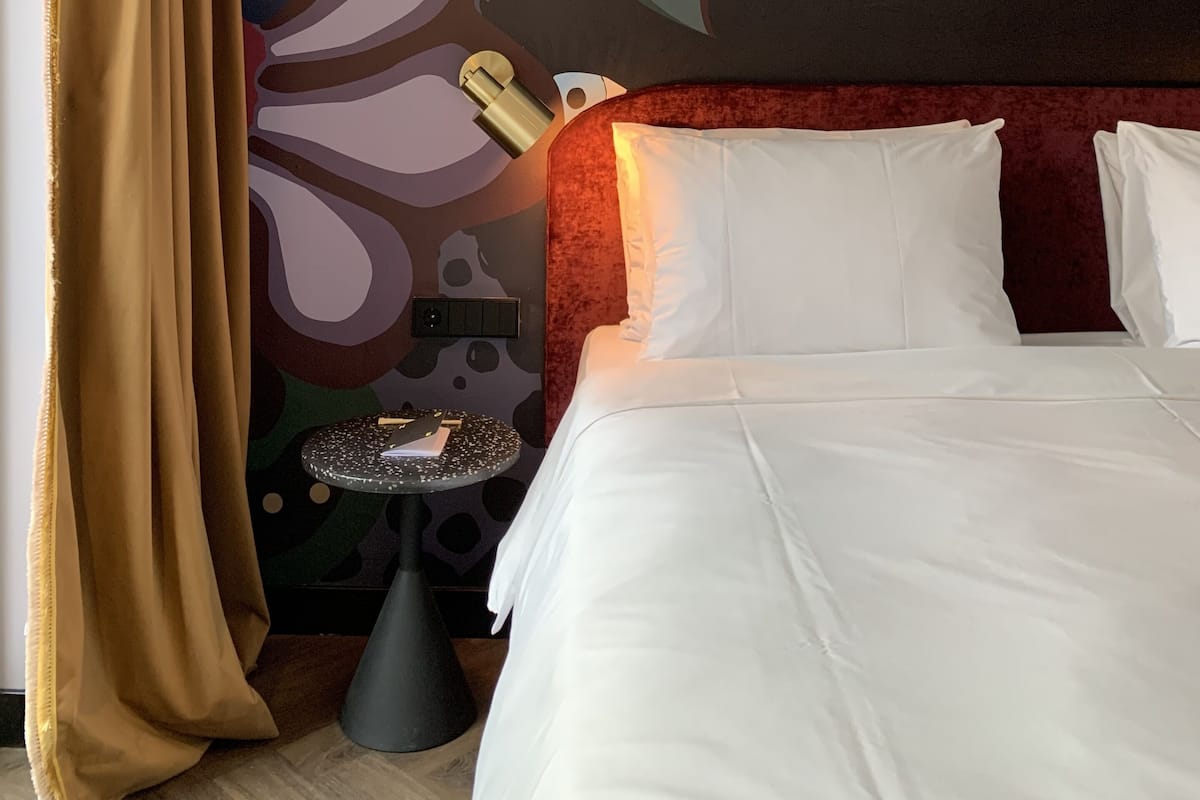 A hotel room with a large bed, a red headboard, a wall with a floral mural, a small round table with a lamp, and a curtain.