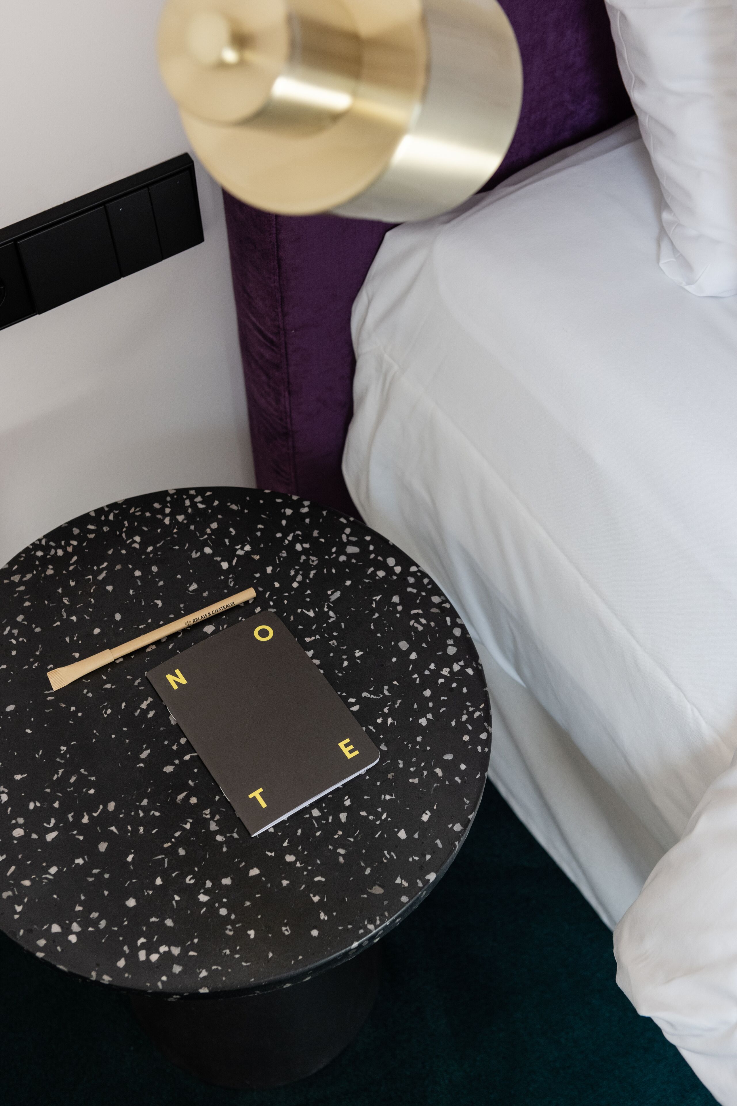 A round black ottoman with a black and gold tray, a black and gold lamp, and a white pillow on a bed with purple bedding.
