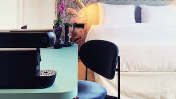Quarto de hotel com cama, escrivaninha com computador, cadeira, vaso com flores e luminária de parede.