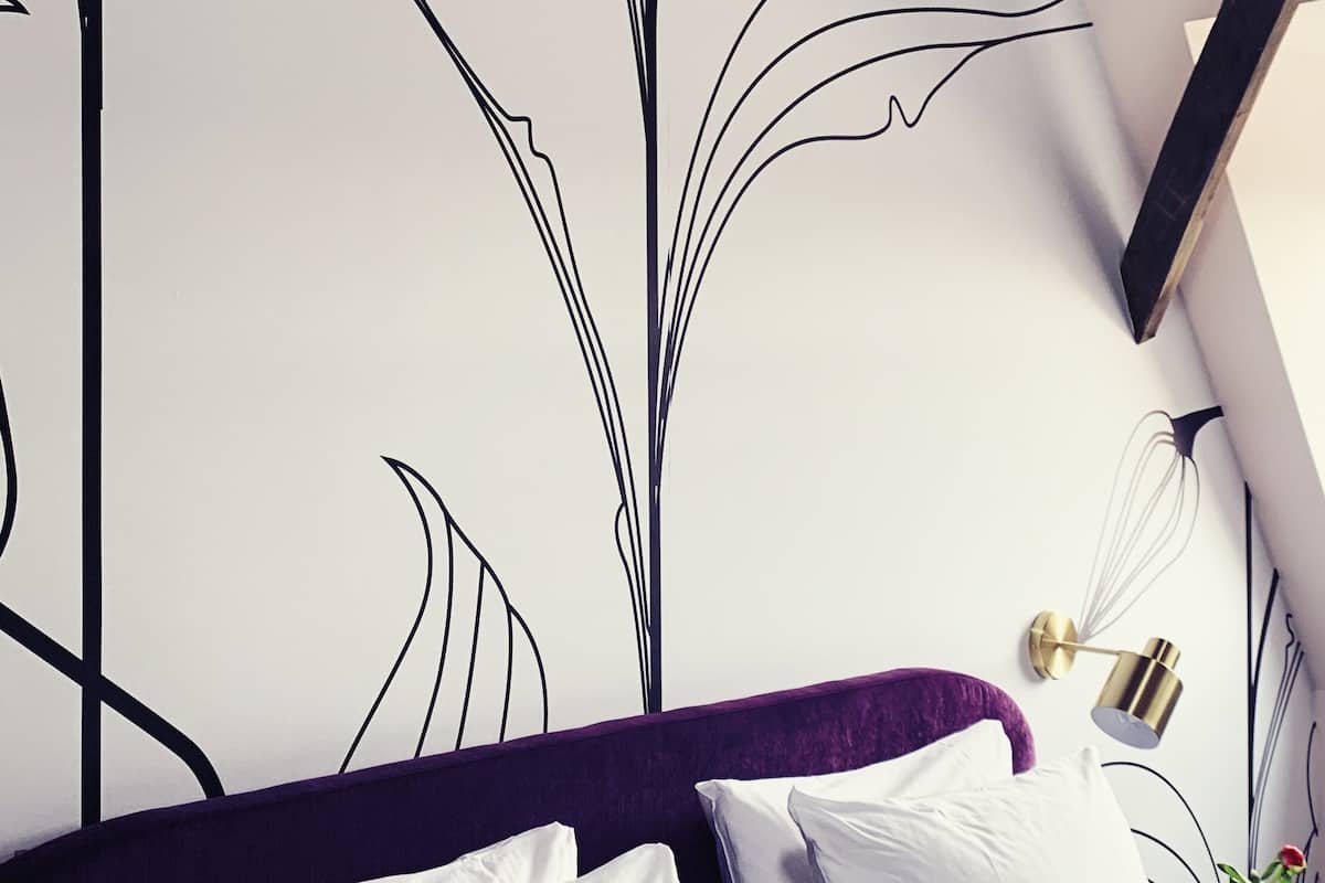 A bedroom with a white bed, purple headboard, and a wall with a black abstract design.