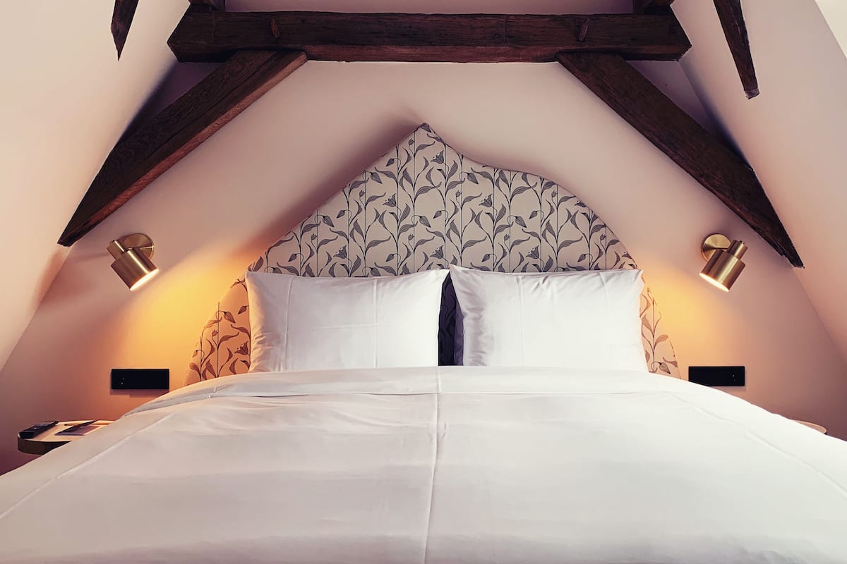 A bedroom with a high, sloped ceiling and exposed wooden beams. There is a bed with white linens and a floral-patterned headboard. Two wall-mounted lamps provide lighting.
