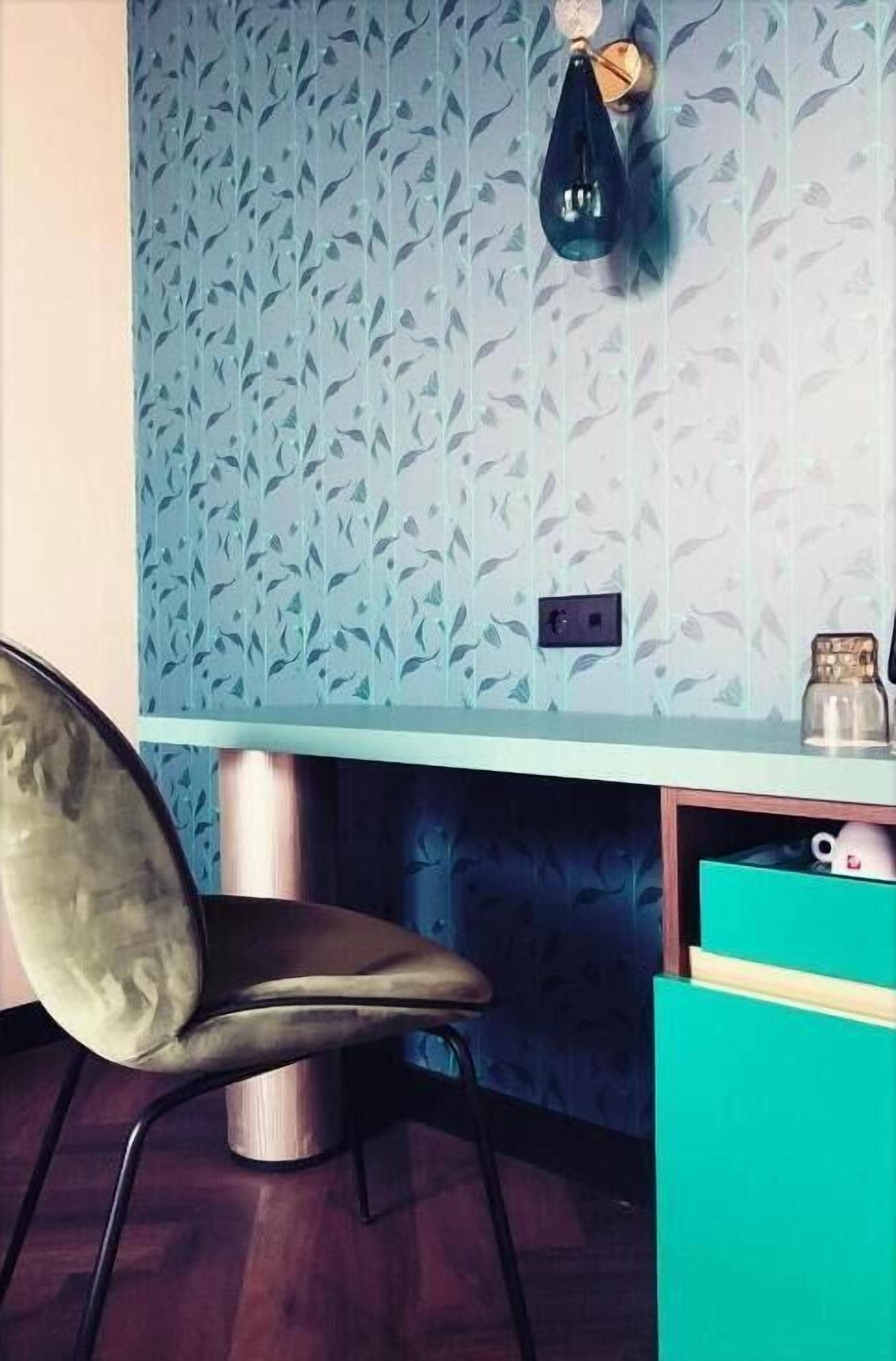 Ein Zimmer mit gemustertem Wandpapier, einem Schreibtisch mit einer daran hängenden Tasche, einem Stuhl und einem türkisblauen Schrank.