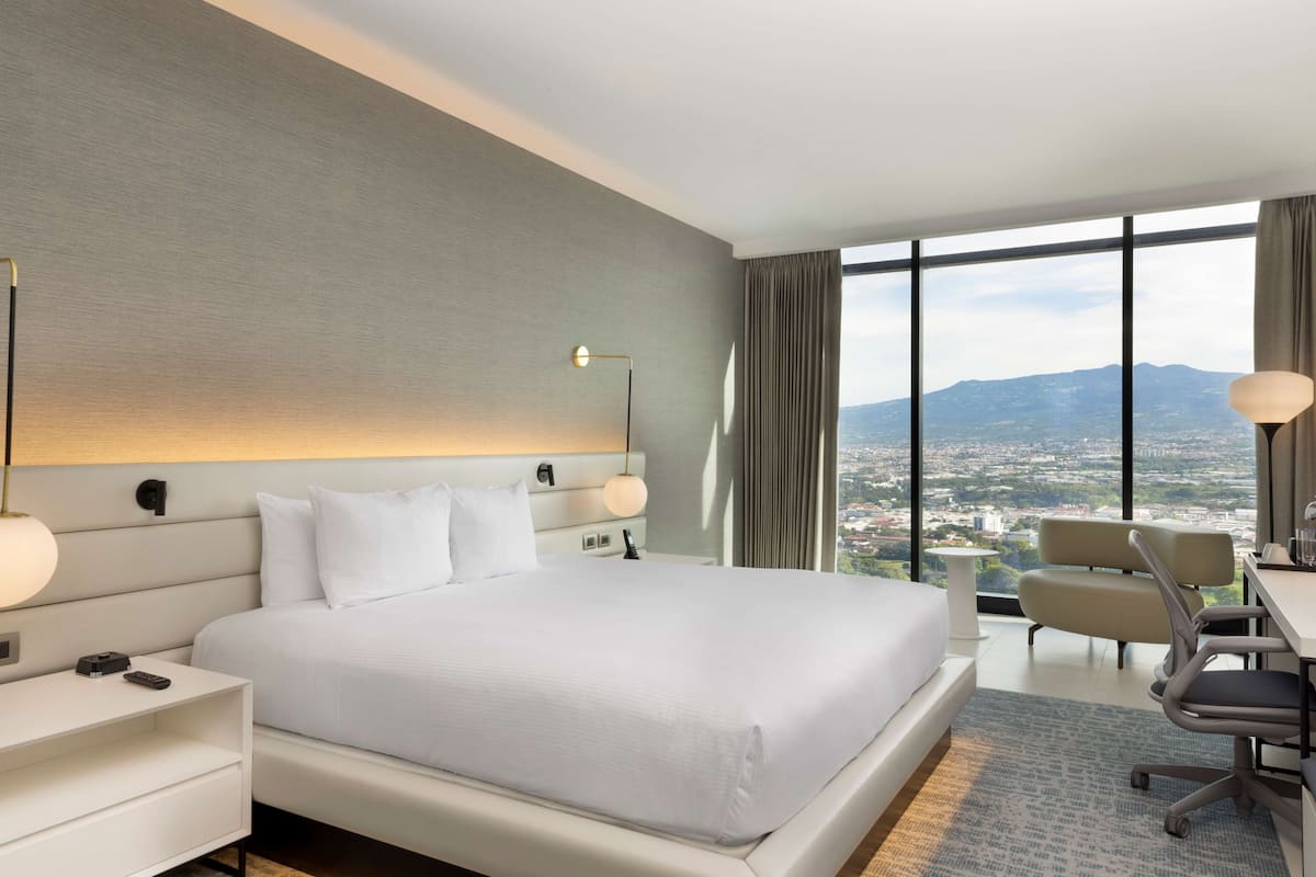 Una habitación de hotel moderna con una cama grande, un escritorio y vistas a un paisaje urbano a través de amplios ventanales.