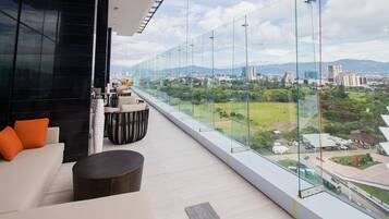 Quarto de hotel moderno com varanda grande, sofá, mesa redonda e vista para a cidade e áreas verdes.