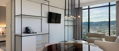 A modern living room with a round glass table, white sofas, a mounted TV, and a city view through large windows.