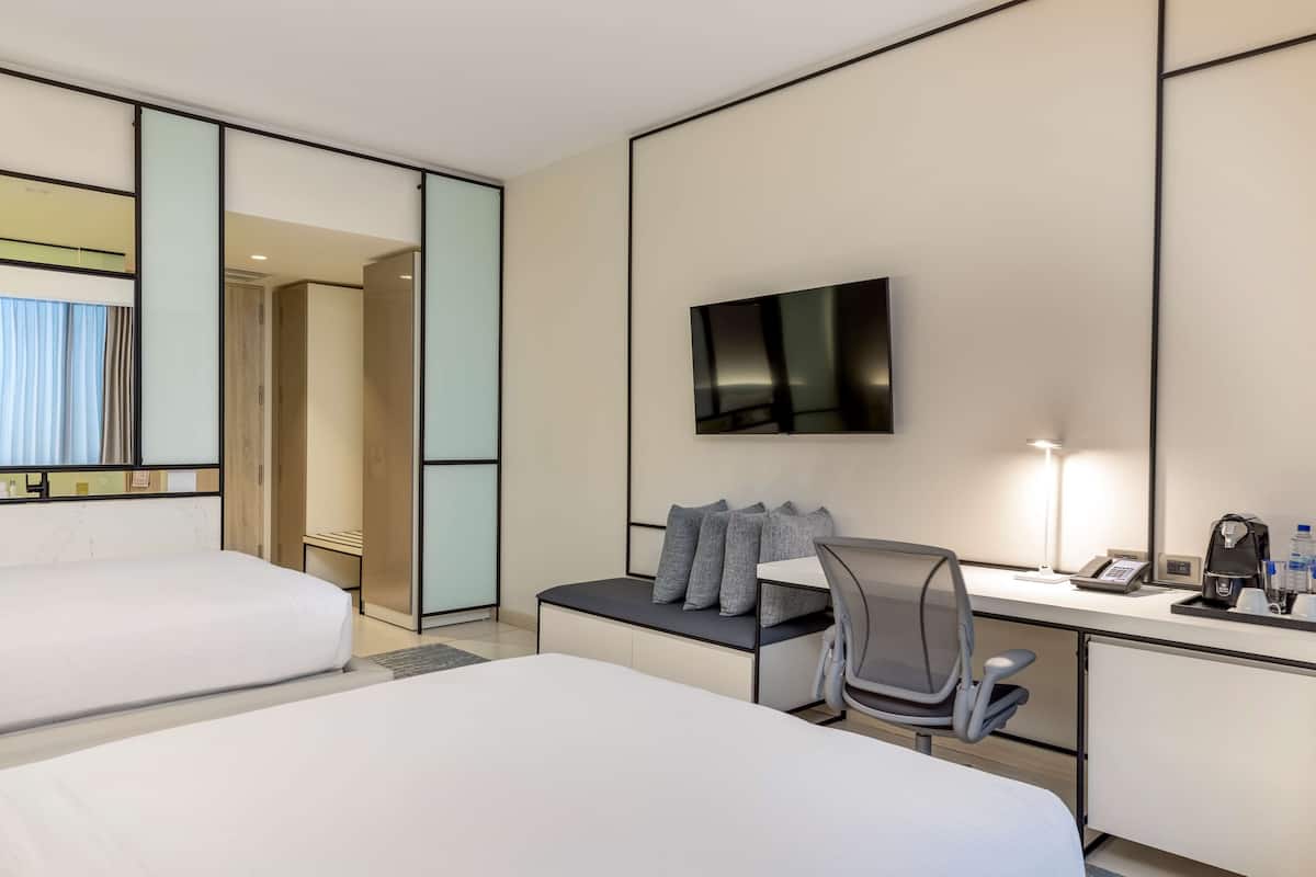 Habitación de hotel con dos camas, un escritorio con silla, televisor de pantalla plana y mesita de noche con lámpara y teléfono.