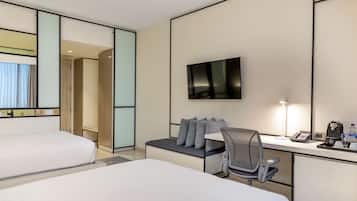 A hotel room with two beds, a desk with a chair, a flat-screen TV, and a bedside table with a lamp and a phone.
