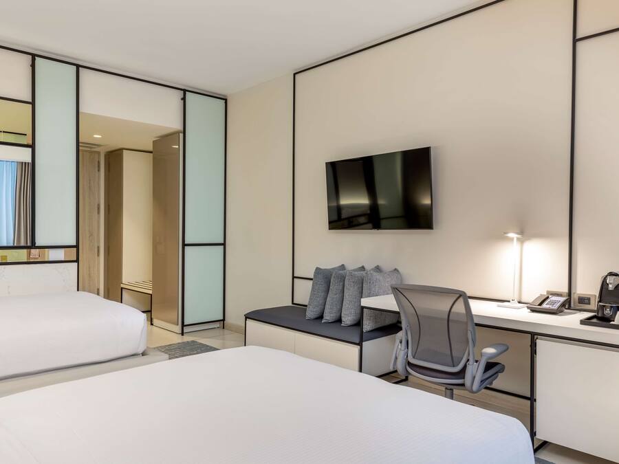 A hotel room with two beds, a desk with a chair, a flat-screen TV, and a bedside table with a lamp and a phone.