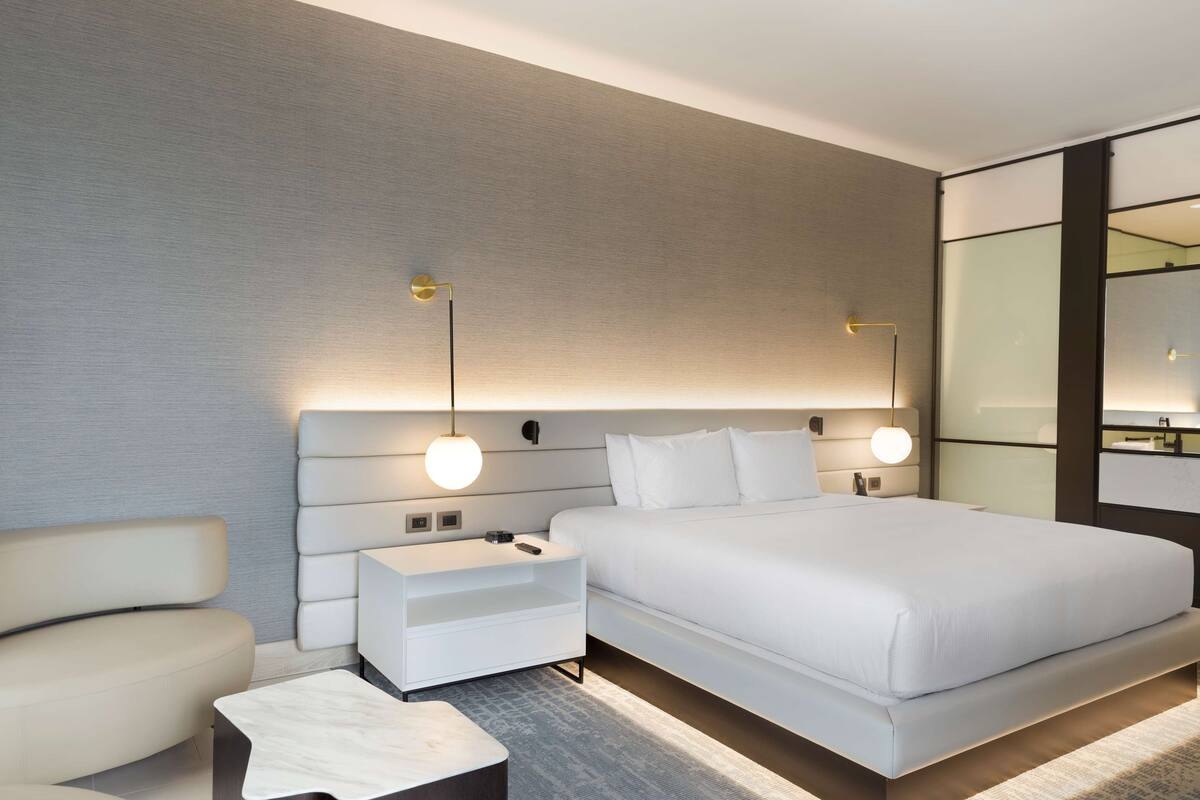 Una habitación de hotel moderna con una cama grande, una mesita de noche, una silla y una mesita auxiliar.