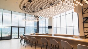 A modern bar area with a curved counter, bar stools, and a large window offering a city view. - Hilton San Jose La Sabana (San José)