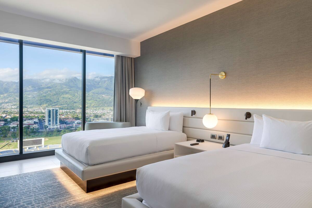 Habitación de hotel con dos camas, un ventanal grande con vistas a la ciudad, una lámpara moderna y una mesita de noche con teléfono.
