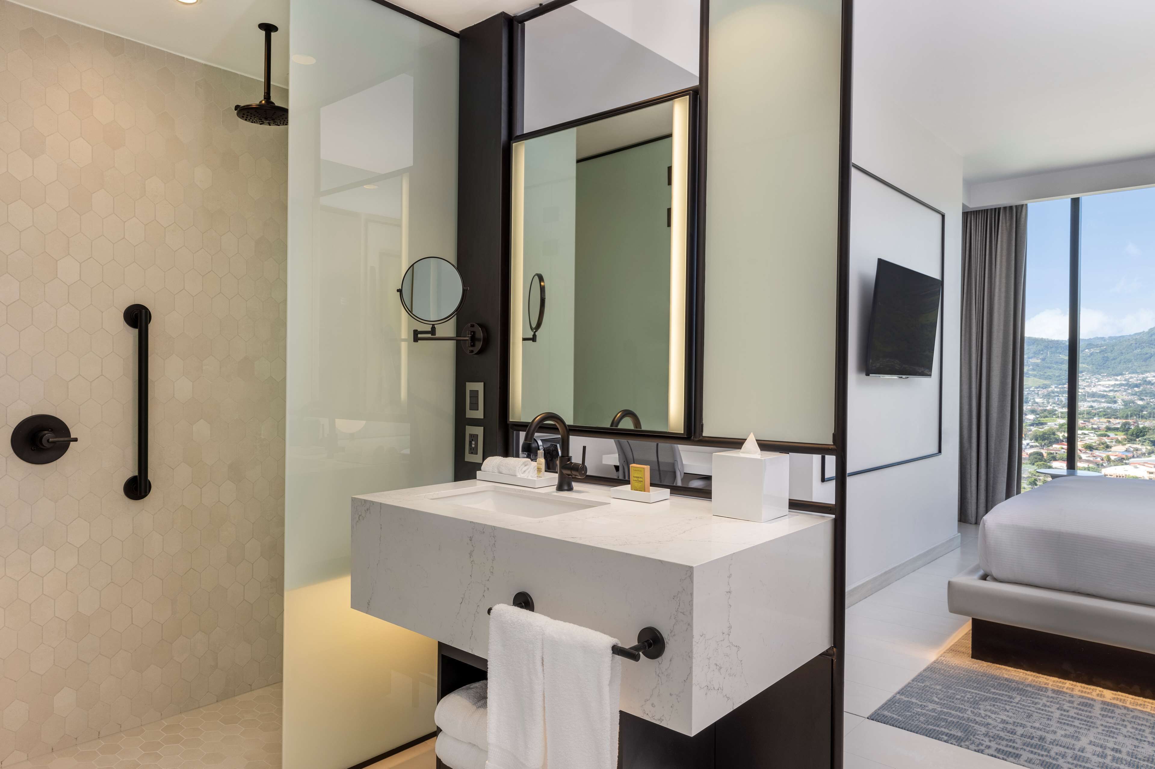 A modern bathroom with a large mirror, two sinks, a shower, and a bed with a city view.