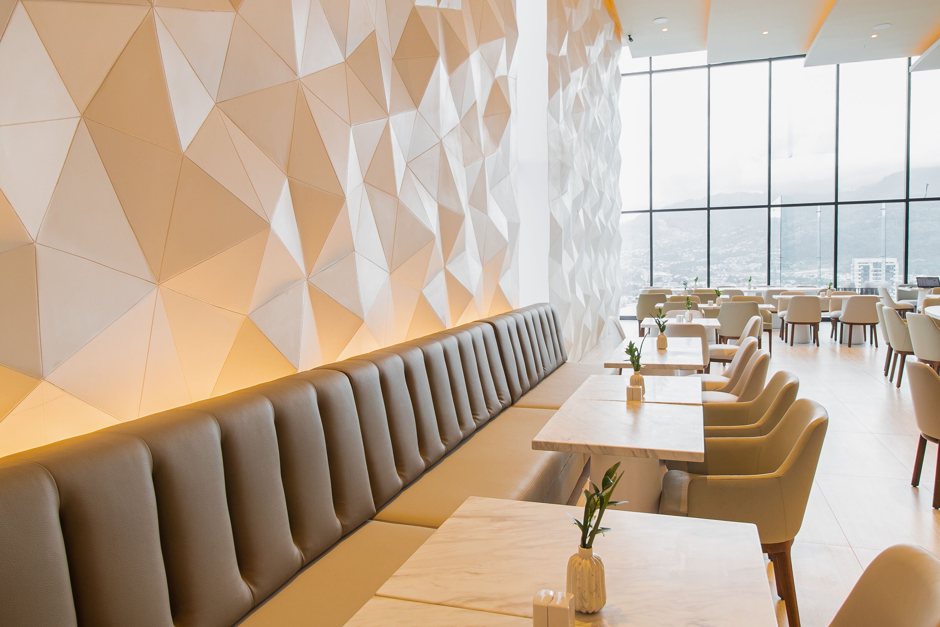 A modern restaurant with a geometric wall design, a long bench seating area, and a view of the city through large windows.