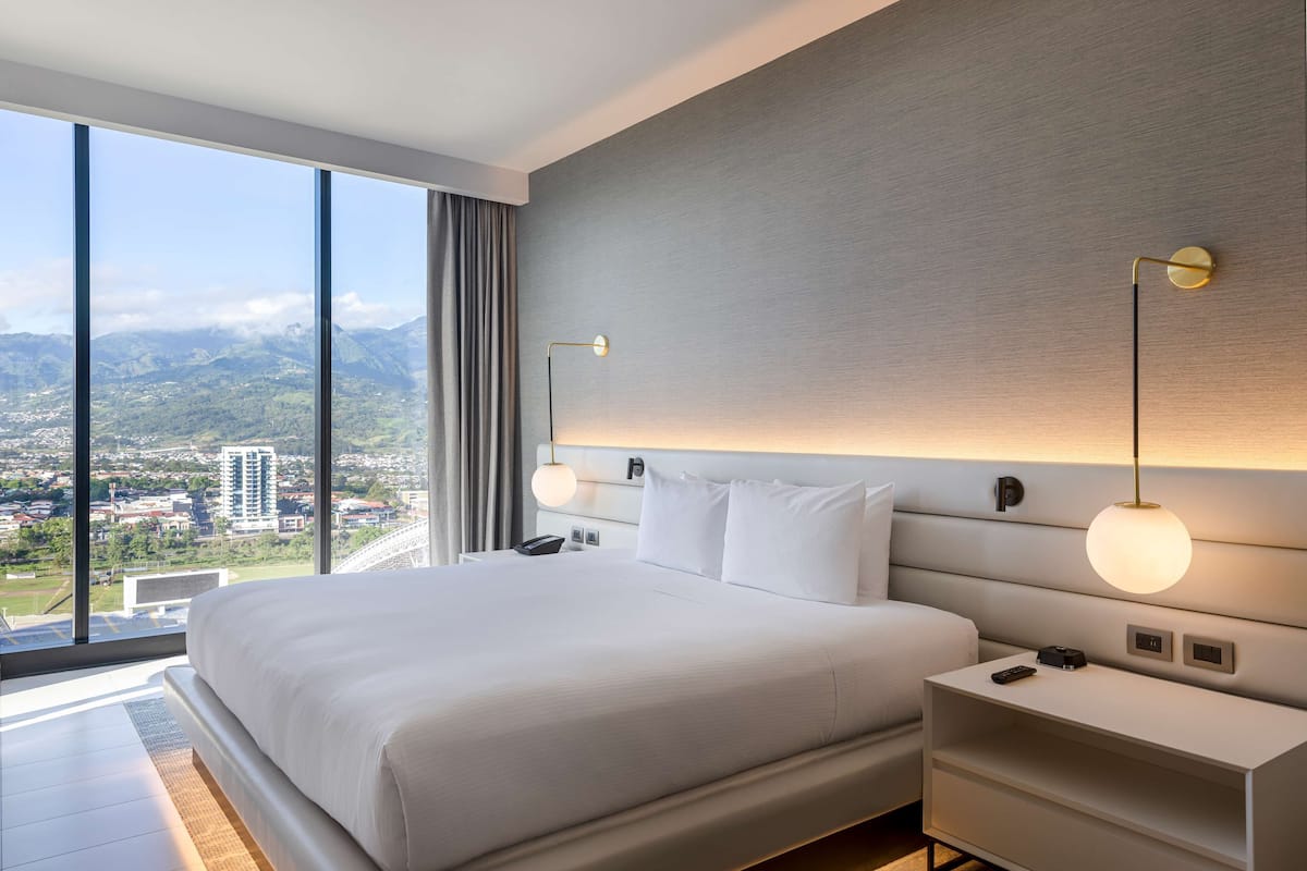 Una habitación de hotel moderna con una cama grande, mesitas de noche y vistas a una ciudad y montañas a través de amplios ventanales.
