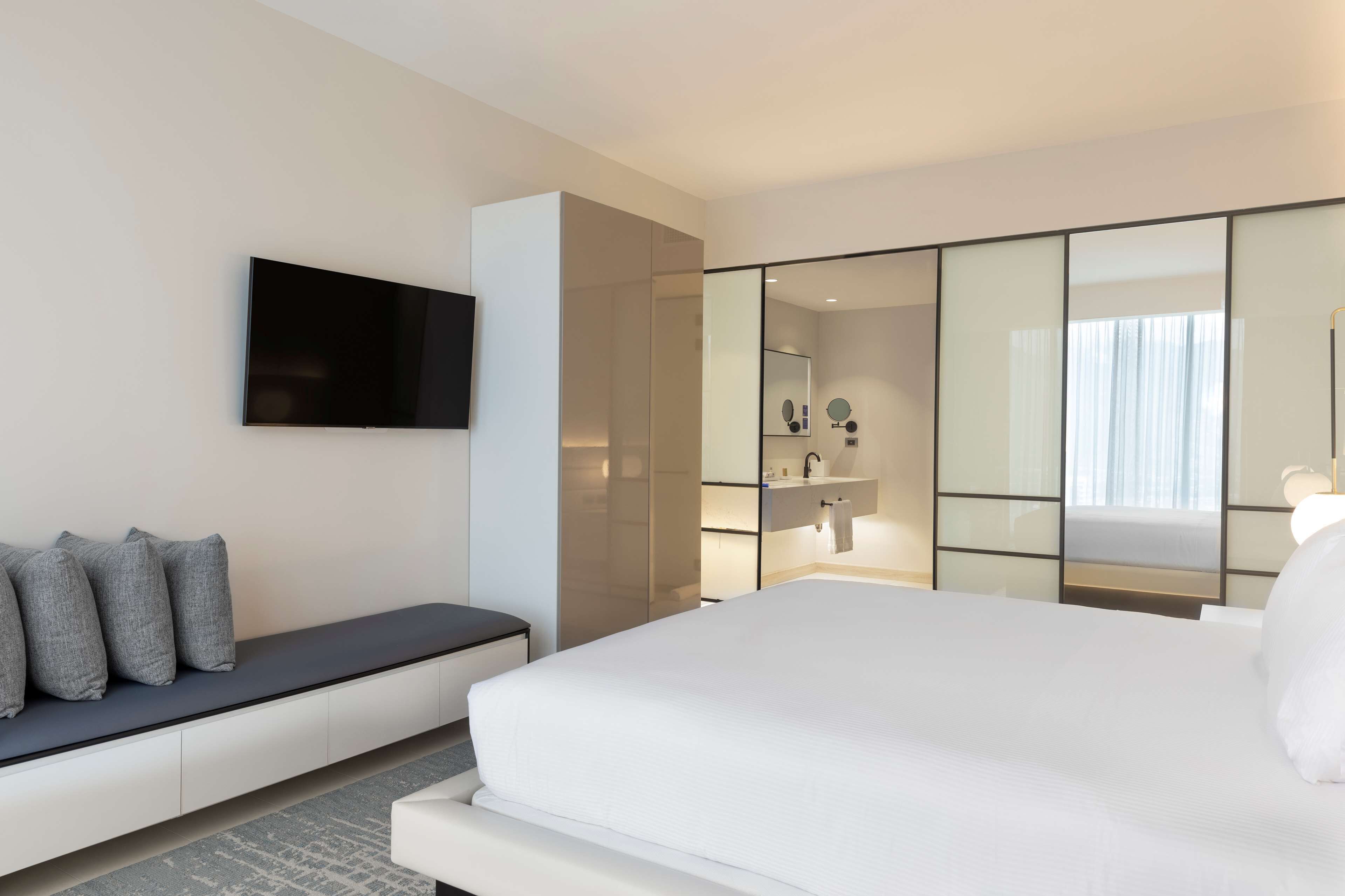 A modern hotel room with a large bed, a seating area with cushions, a flat-screen TV mounted on the wall, and a large mirror reflecting the room.