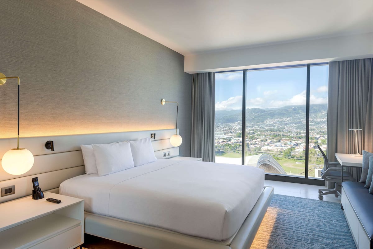 Habitación de hotel moderna con una cama grande, un escritorio y vistas a un paisaje urbano a través de amplias ventanas.