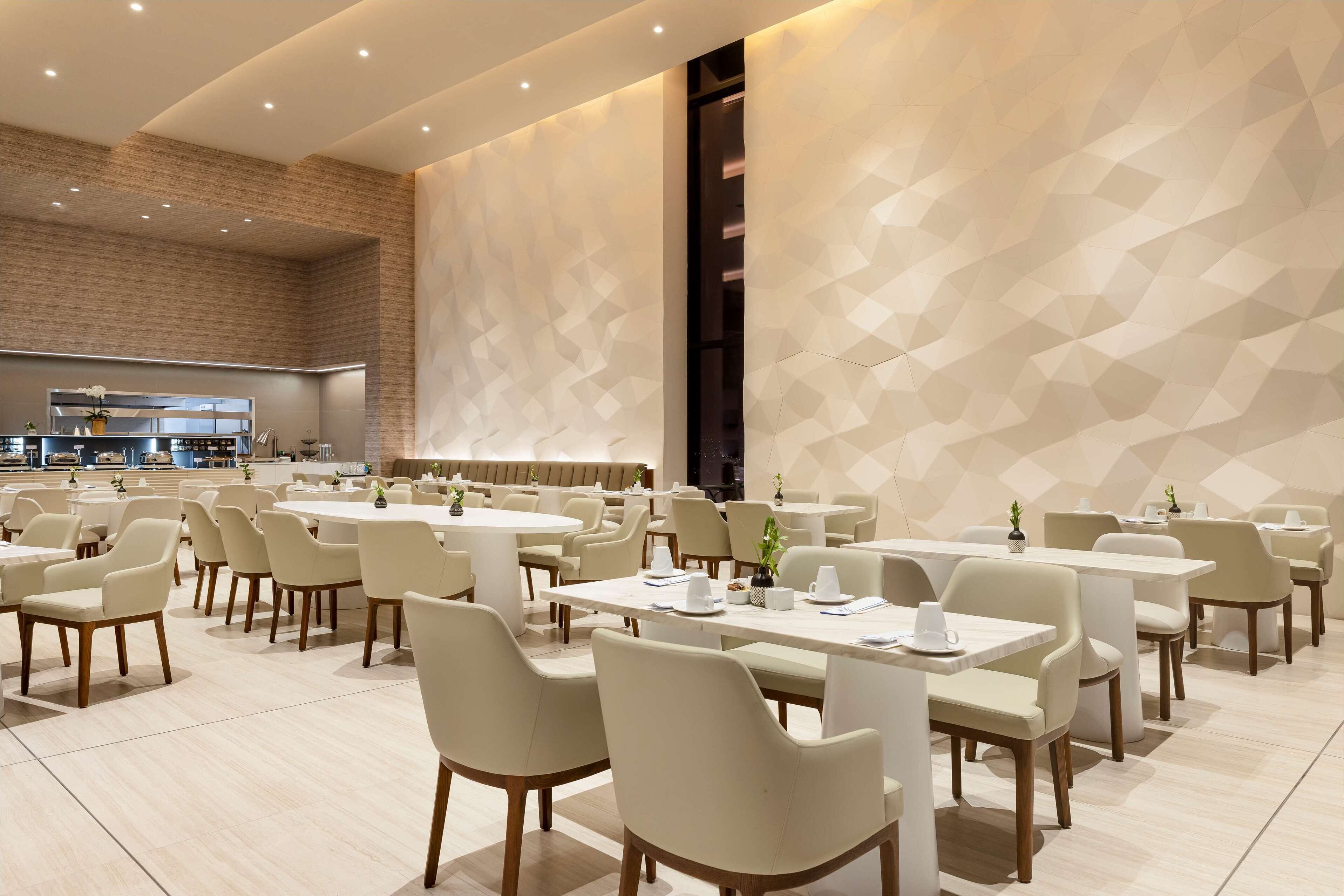 A spacious dining area with multiple tables and chairs, a kitchen in the background, and a geometric patterned wall.