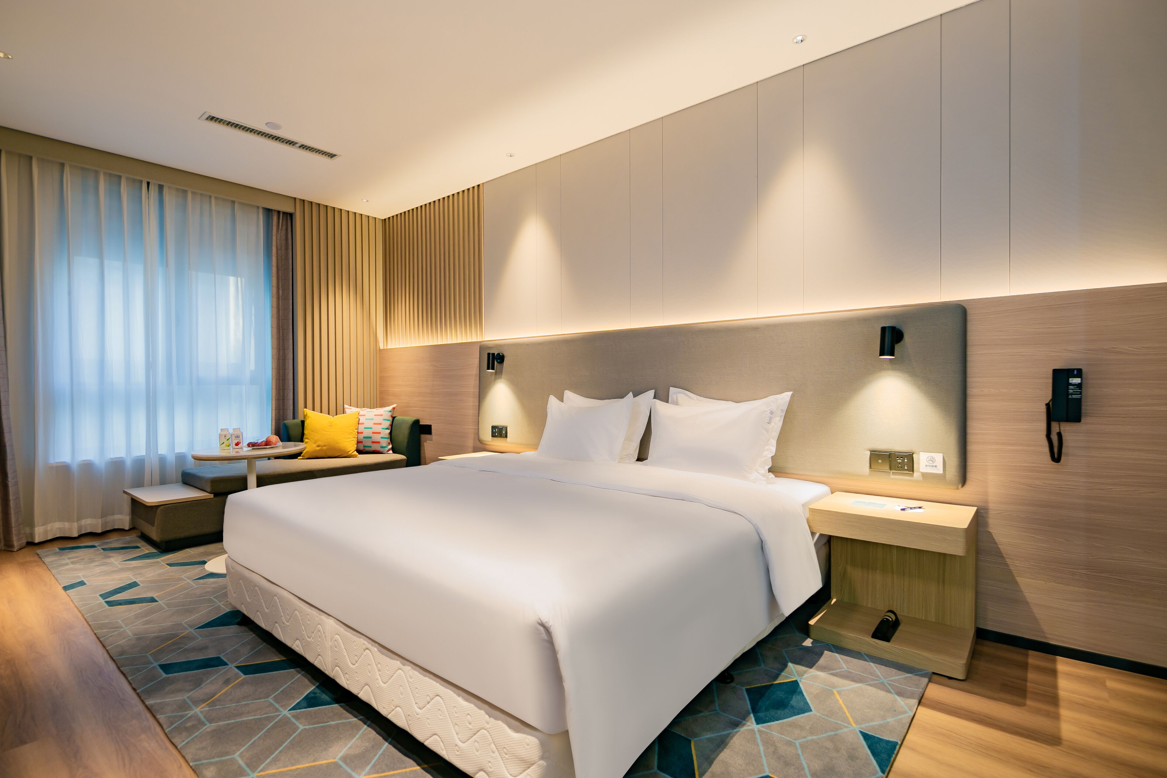 A modern hotel room with a large bed, bedside tables, a nightstand, and a wall-mounted phone.
