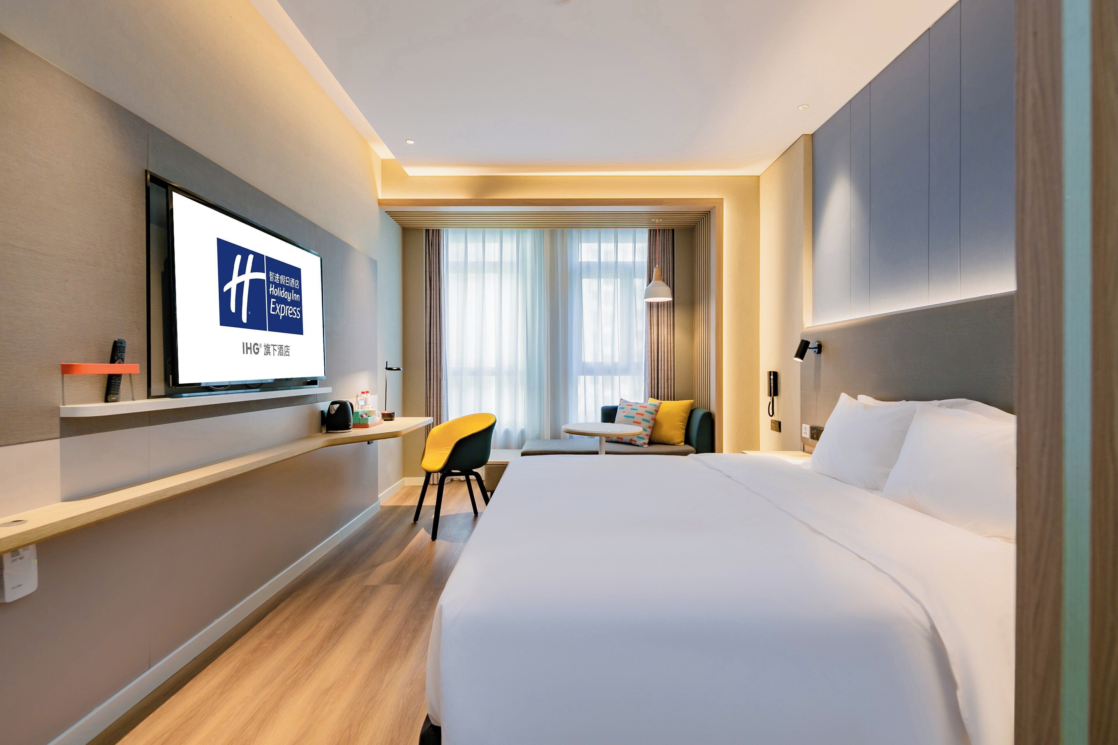Hotelkamer met een bed, een bureau, een stoel, een aan de muur bevestigde televisie en een raam met gordijnen.