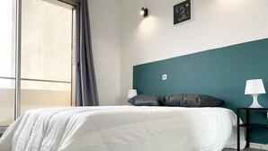Een netjes opgemaakt bed met wit beddengoed, een wand in teal, een klein nachtkastje met een lamp, en een raam met gordijnen.
