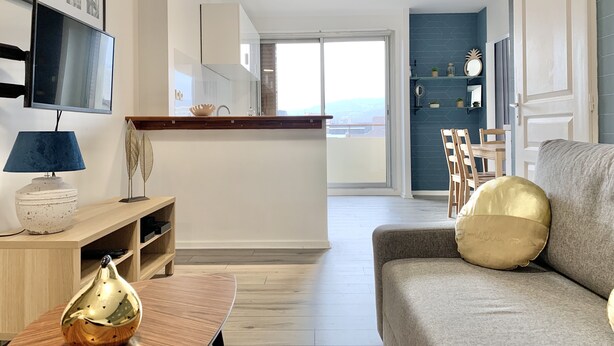 Een moderne woonkamer met een bank, een salontafel en een televisie. Er is een keuken met een spoelbak en kastjes, en een eethoek met stoelen.