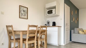Een ruimtelijk ingerichte leefruimte met een houten eettafel en stoelen, een wit kastje met een magnetron, koffiezetapparaat en decoratieve wandkunst.