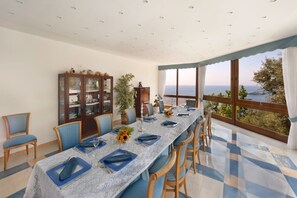 Un comedor con una mesa larga dispuesta para seis personas, rodeado de sillas azules, un aparador de cristal con adornos y vistas al mar a través de amplios ventanales.