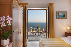 Habitación de hotel con cama, mesita de noche, orquídea en maceta, balcón con mesa y sillas, y un cuadro en la pared.