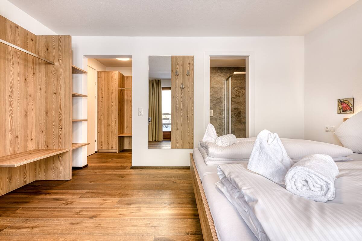 Camera d'albergo con pavimento in legno, un letto grande con lenzuola bianche, un armadio in legno e zona doccia.