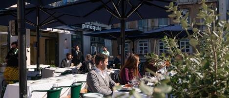 Un área de comedor al aire libre con mesas dispuestas bajo sombrillas, un letrero de restaurante visible y un edificio en el fondo.
