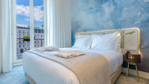 Habitación de hotel con una cama grande, una mesita de noche con lámpara y vistas a edificios a través de una ventana.