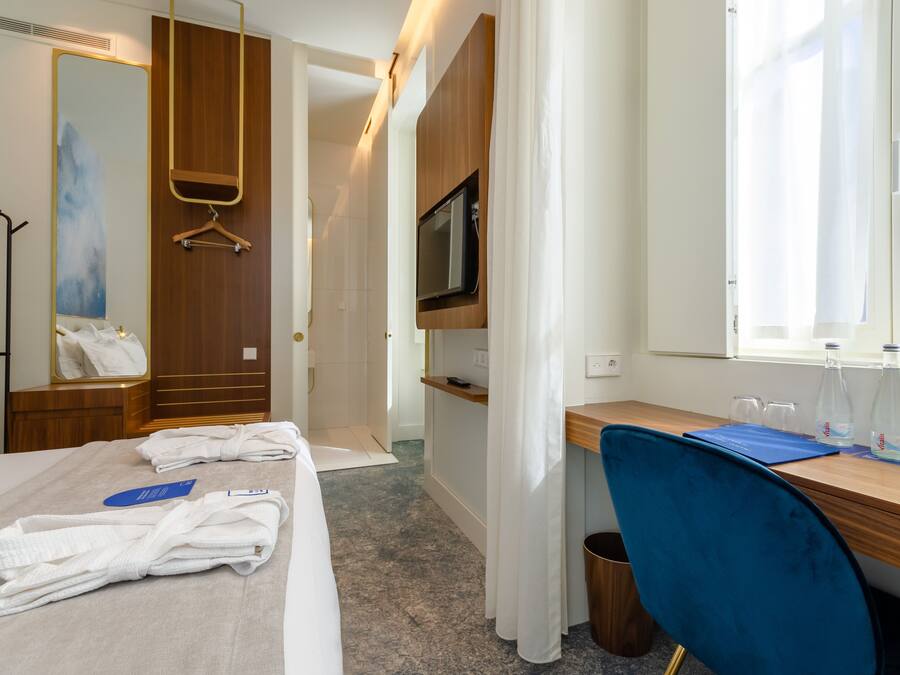 Ein Hotelzimmer mit einem Bett, einem Schreibtisch mit Stuhl, einem Fernseher, einem Spiegel und einem Fenster mit Vorhängen.