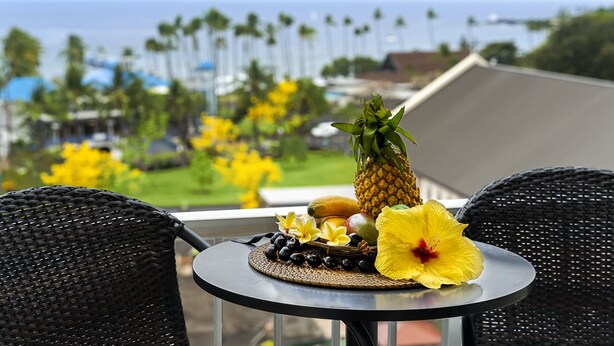 A table with a tropical fruit arrangement, including a pineapple and a yellow flower, on a balcony overlooking a coastal area with palm trees. - Kailua Village #701: Top Floor, Corner Penthouse Remodeled in Downtown Kona. (Kailua Kona)