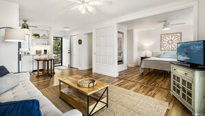 A spacious living room with a wooden coffee table, a sofa, a TV, and a ceiling fan. - Kailua Village #701: Top Floor, Corner Penthouse Remodeled in Downtown Kona. (Kailua Kona)