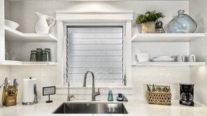 A modern kitchen with a stainless steel sink, white cabinetry, and a window with horizontal blinds. - Kailua Village #701: Top Floor, Corner Penthouse Remodeled in Downtown Kona. (Kailua Kona)