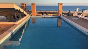 Una piscina infinita con vistas al océano, una terraza con sol y sillones de descanso, y una pequeña construcción de ladrillo.