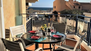 Mesa redonda con sillas en un balcón con vistas al mar, una botella de champán y un jarrón de flores.