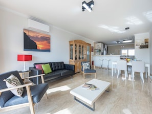 A modern living room with a sofa, chairs, a coffee table, a cabinet, and a kitchen area with a bar. - Mira Baia - Meia Praia Family Apartment (Lagos)