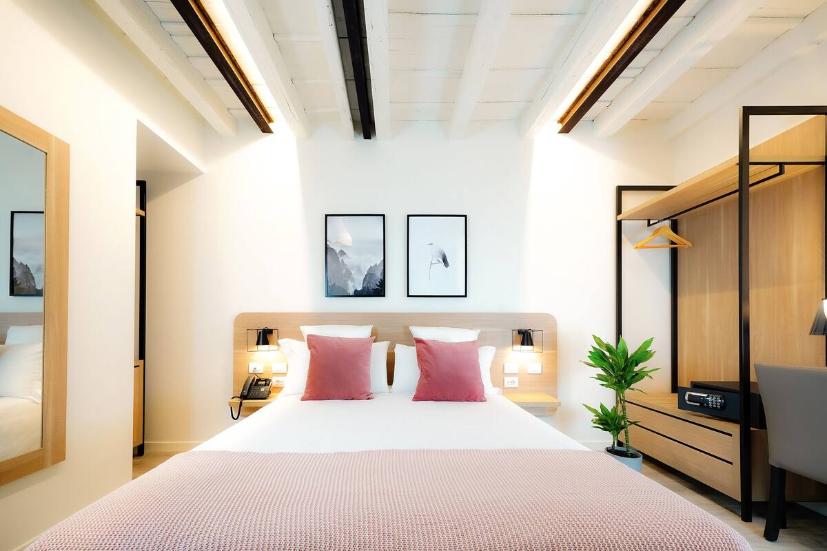 A modern hotel room with a large bed, bedside lamps, a desk with a chair, a hanging plant, and two framed pictures on the wall.