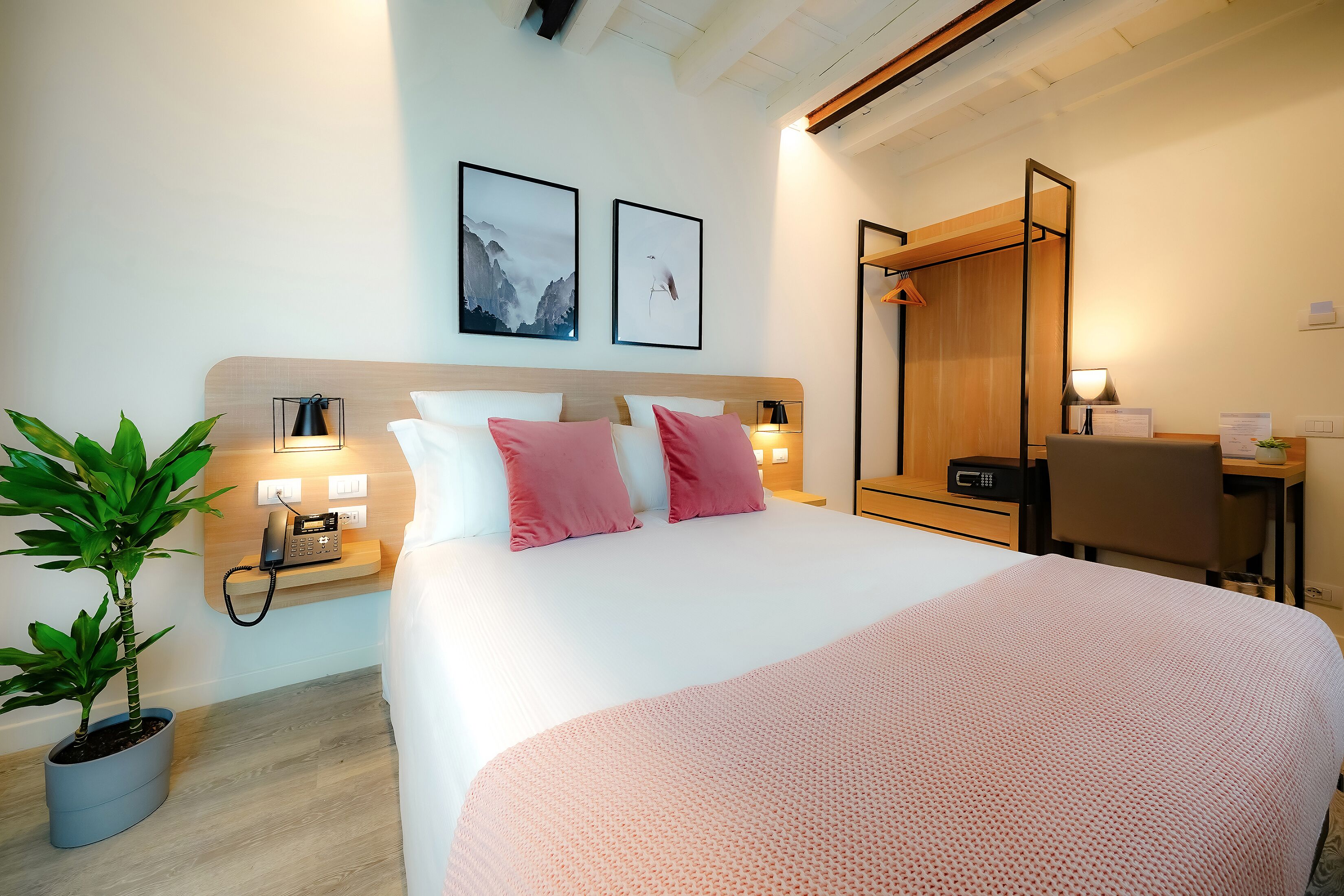 A hotel room with a large bed, a desk, a chair, a potted plant, and artwork on the wall.