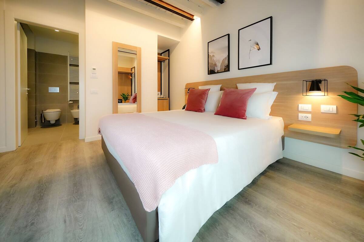 A modern bedroom with a large bed, bedside tables, a nightstand, and a wall-mounted lamp.