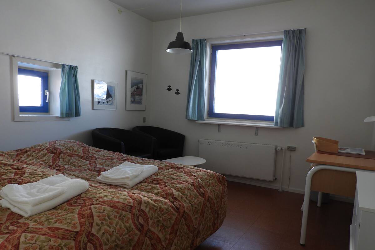 A bedroom with a bed, a desk, a chair, a window with curtains, and a radiator.