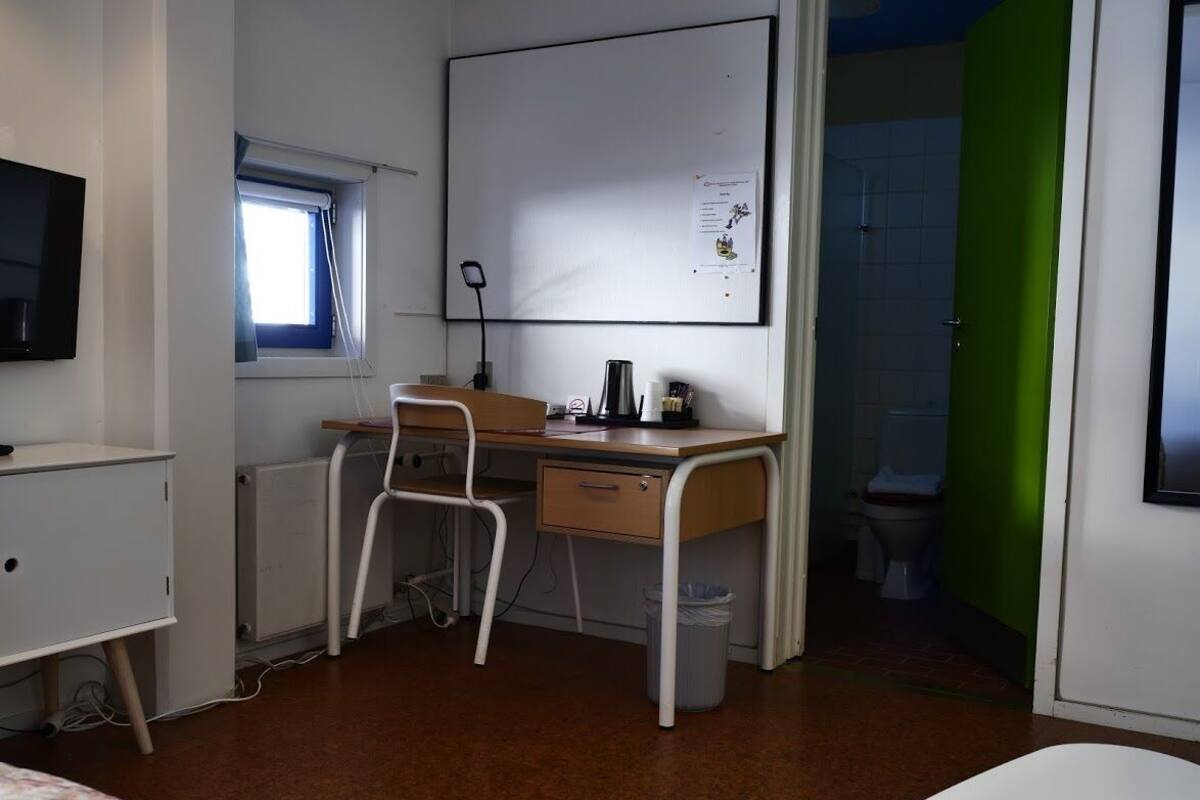 A small, functional room with a desk, chair, and a bed. There is a window with blinds, a whiteboard, and a green door.