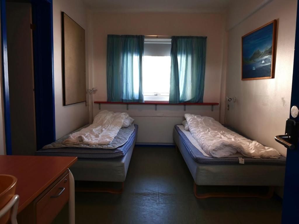 A small room with two beds, a window with curtains, a desk, and a painting on the wall.
