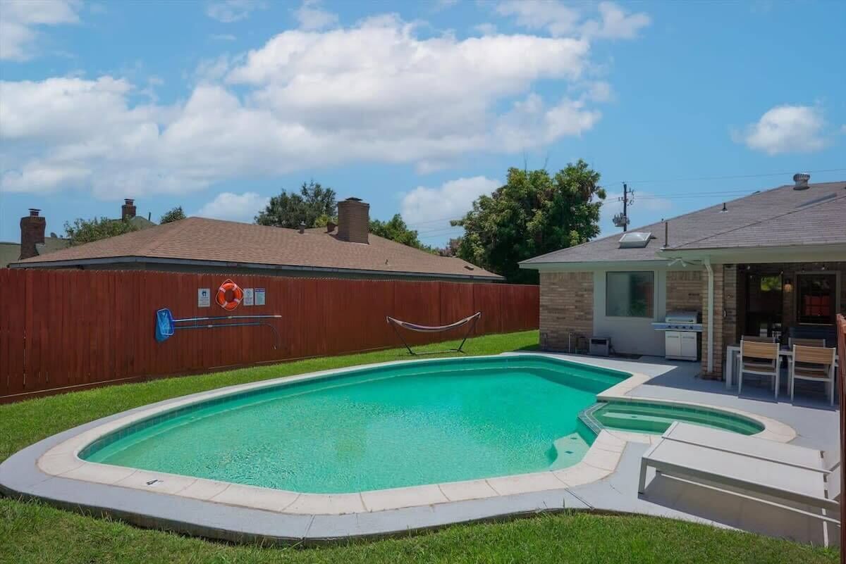 En swimmingpool med ett trästaket, en täckt uteplats och en byggnad i tegel som syns i bakgrunden.