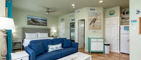 Una habitación con una cama, un sofá azul, una mesa de centro blanca, un ventilador de techo y una cocina con armarios y un refrigerador.