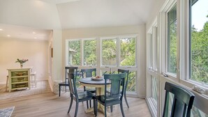 A dining area with a round table and chairs, large windows with a view of greenery, and a light-colored floor.